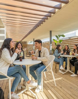 Étudiants assis en terrasse sous une pergola en bois, discutant et buvant des rafraîchissements dans un espace de détente lumineux